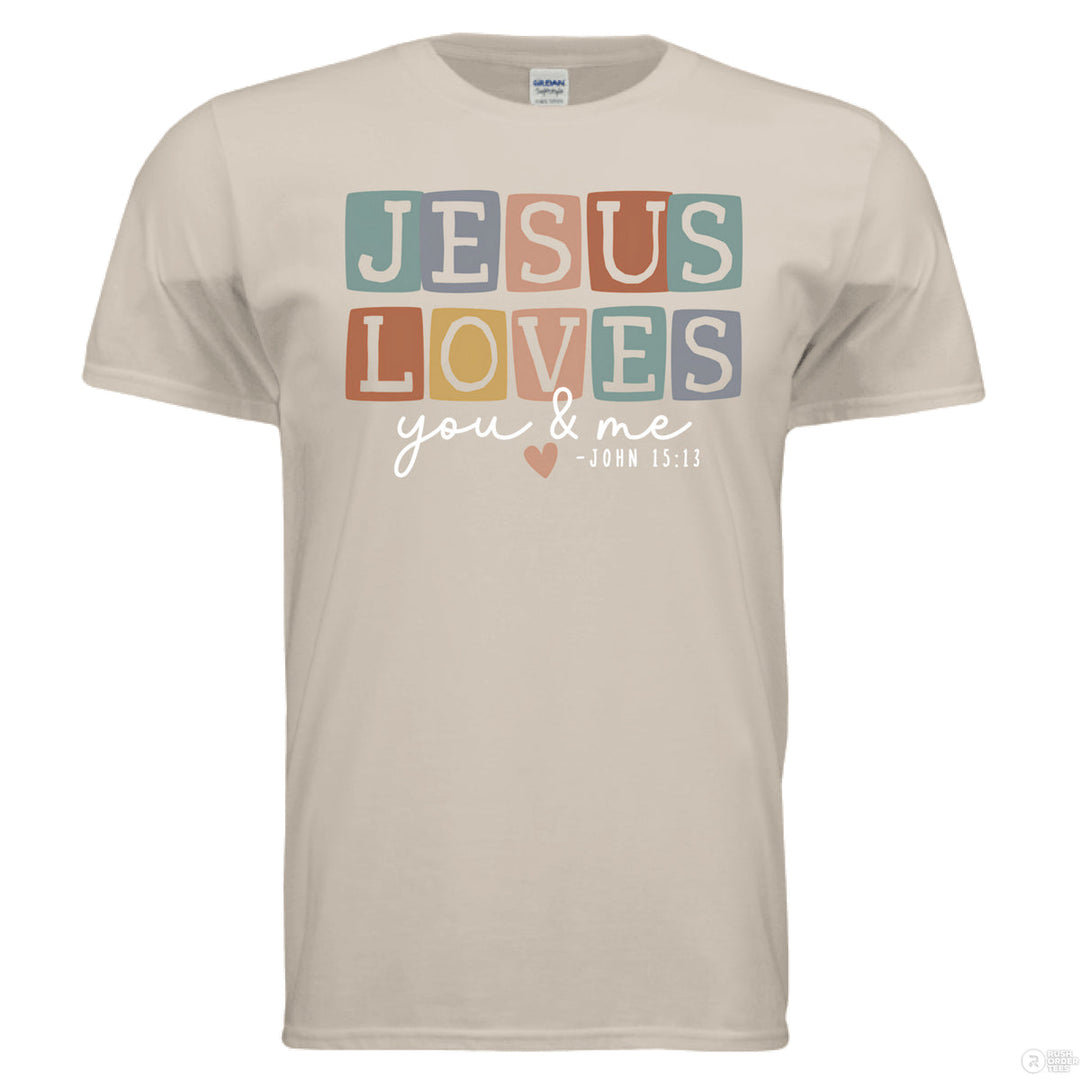 Sand shirt with pastel block letters reading “Jesus Loves You & Me” and a heart, referencing John 15:13.
