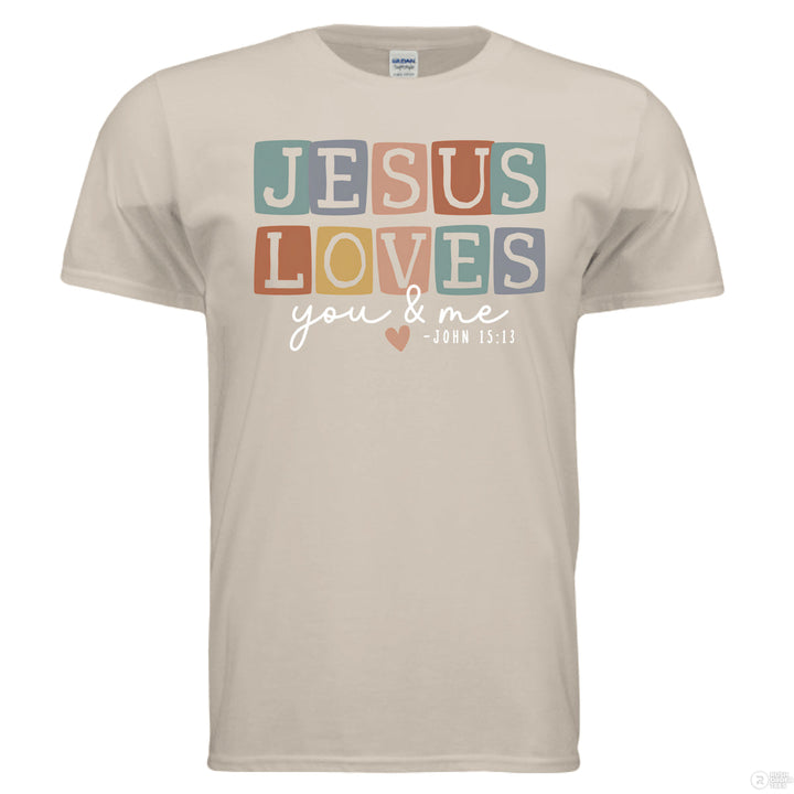 Sand shirt with pastel block letters reading “Jesus Loves You & Me” and a heart, referencing John 15:13.